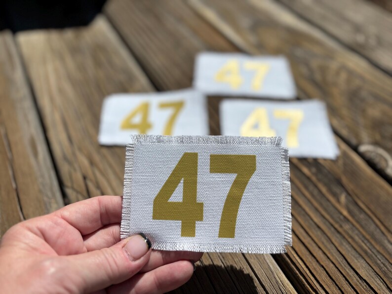 May include: A hand holds a rectangular patch with the number 47 printed in gold. The patch is white with a frayed edge. Three other patches with the number 47 are in the background.