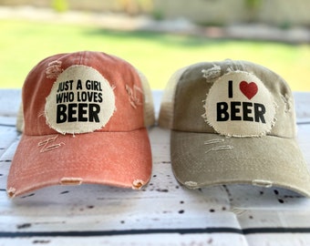 Puede incluir: Dos gorras de béisbol desgastadas con parches. Una gorra es roja con un parche blanco que dice "Just a girl who loves beer". La otra gorra es beige con un parche blanco que dice "I love beer" con un corazón rojo.