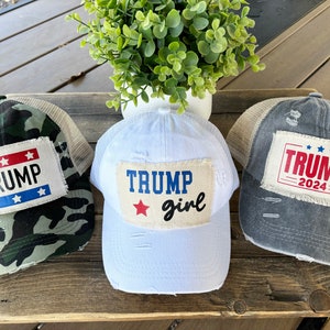 May include: Three trucker hats with distressed fabric and patches. The hats are in camouflage, white, and grey. The patches feature the text "TRUMP" with stars and the year "2024".