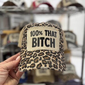 May include: A leopard print baseball cap featuring a rectangular beige patch on the front. The patch has frayed edges and displays the black text 100% THAT BITCH. The cap has a structured crown and a curved brim with a distressed, worn-in appearance.