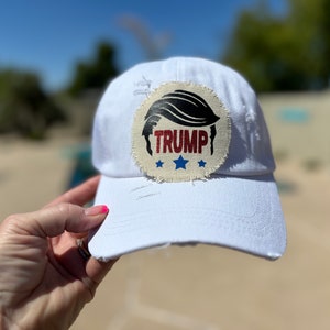Może przedstawiać: Biała czapka bejsbolówka z przetartym naszywką z tkaniny przedstawiającą czarną sylwetkę włosów i napis "TRUMP" czerwonymi literami z dwoma niebieskimi gwiazdami.