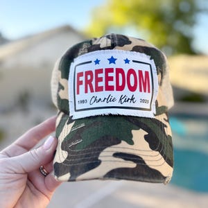 May include: Camouflage baseball cap with a white patch that reads "FREEDOM" in red letters, with three blue stars above the word. The patch also includes the text "1993 Charlie Kirk 2025". The cap has a mesh back.