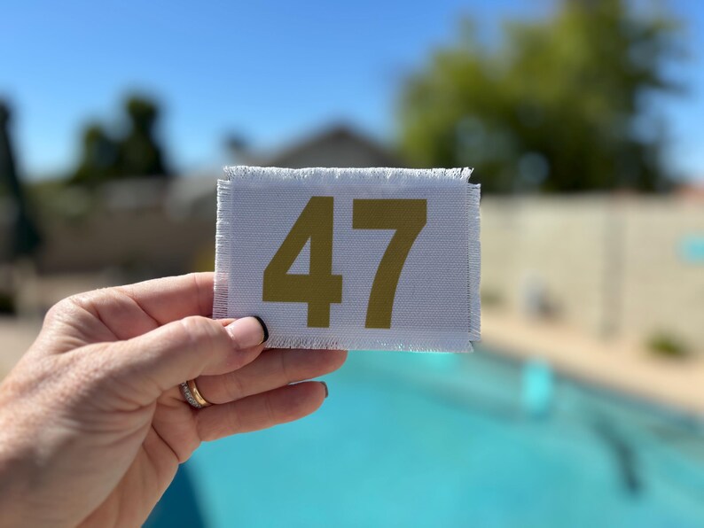 May include: A white rectangular patch with frayed edges, displaying the number 47 in gold. The patch is held against a blurred background of a swimming pool and blue sky.