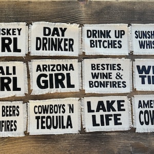 May include: Twelve white fabric patches with black text. The patches have frayed edges and feature phrases like "Whiskey Girl", "Day Drinker", "Cali Girl", "Arizona Girl", "Boots, Beers 'N Bonfires", "Cowboys 'N Tequila", "Drink Up Bitches", "Sunshine & Whiskey", "Besties, Wine & Bonfires", "Wine Time", "Lake Life", and "American Cowgirl".