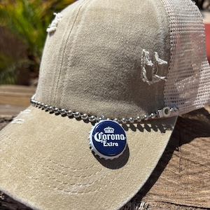 May include: Tan trucker hat with a distressed look, featuring a white mesh back and a silver ball chain. A Corona Extra bottle cap charm hangs from the chain, adding a unique touch to this casual accessory.