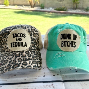 Może przedstawiać: Dwie czapki bejsbolowe. Jedna ma nadruk w cętki z tekstem "TACOS AND TEQUILA" na beżowej naszywce. Druga jest turkusowa z tekstem "DRINK UP BITCHES" na beżowej naszywce. Obie mają siatkowy tył.