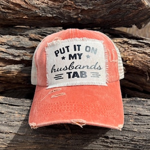 Puede incluir: Una gorra de béisbol color coral desgastada con parte trasera de malla blanca y un parche rectangular en el frente. El parche tiene bordes deshilachados y el texto negro: PUT IT ON MY husbands TAB, decorado con pequeñas estrellas y líneas.
