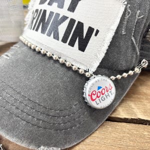 May include: Gray trucker hat with a mesh back and a distressed brim. The hat features a patch with the words "DAY DRINKIN'" and a Coors Light bottle cap charm attached with a beaded chain.
