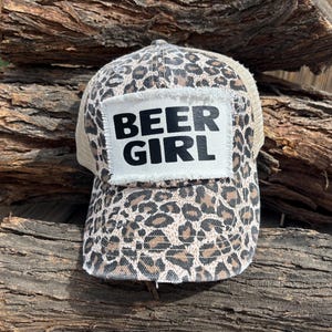 Może przedstawiać: Czapka typu trucker z nadrukiem w panterkę i białą naszywką z napisem "BEER GIRL" czarnymi literami. Czapka ma beżowy tył z siatki i zakrzywiony daszek.