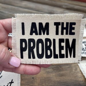 May include: A rectangular, beige fabric patch with frayed edges. Bold, black text reads "I AM THE PROBLEM." Other patches with text are visible in the background. The patch is held by a person.