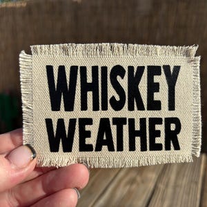 May include: A rectangular patch with frayed edges, featuring the words "WHISKEY WEATHER" in bold black letters on a beige background. The patch is held up by a hand.