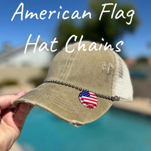 May include: A tan baseball cap with a white mesh back, featuring a chain and an American flag charm. The text "American Flag Hat Chains" is displayed in white script above the cap.