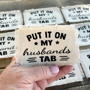 May include: A set of eight white fabric coasters with black text that reads "PUT IT ON MY HUSBANDS TAB". The coasters have a frayed edge and are arranged in a pile.