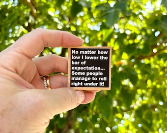 May include: A wooden block with a black background and white text that reads: "No matter how low I lower the bar of expectation... Some people manage to roll right under it!"