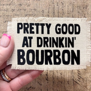 May include: A rectangular off-white fabric patch with frayed edges featuring the black text PRETTY GOOD AT DRINKIN' BOURBON printed in a bold sans-serif font. The patch has a woven texture and is presented as a flat, horizontal piece of material.