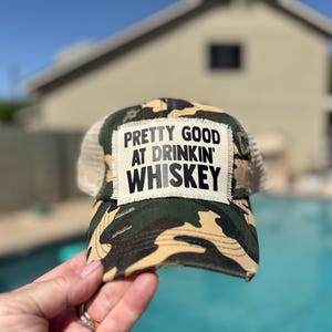 May include: Camouflage trucker hat with a mesh back and a patch that reads "PRETTY GOOD AT DRINKIN' WHISKEY" in black text. The hat is held up by a hand.
