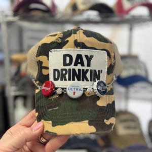 May include: Camouflage trucker hat with the text "DAY DRINKIN'" on a white patch. The hat is decorated with bottle caps and a chain. The hat has a tan mesh back and a camouflage front.