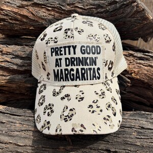 Może przedstawiać: Beżowa czapka typu trucker z wzorem w cętki i siatkowym tyłem. Na przedniej naszywce widnieje napis "PRETTY GOOD AT DRINKIN' MARGARITAS" czarnymi literami. Czapka leży na kawałku drewna.