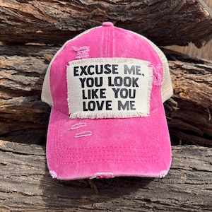 Może przedstawiać: Zniszczona różowa czapka typu trucker z beżowym tyłem z siatki. Na przedniej naszywce widnieje napis "EXCUSE ME, YOU LOOK LIKE YOU LOVE ME" czarnym tekstem. Czapka ma zakrzywiony daszek i zużyty wygląd.