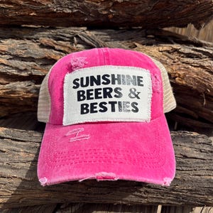 Może przedstawiać: Zniszczona różowa czapka typu trucker z białą naszywką z czarnym napisem "SUNSHINE BEERS & BESTIES". Czapka ma siatkowy tył i zakrzywiony daszek.