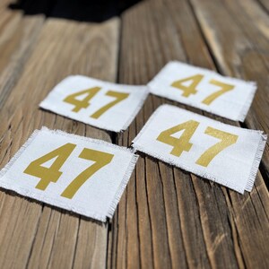 May include: Four white fabric patches, each displaying the number 47 in gold. The patches have a frayed edge and are set against a weathered wooden background. The patches are square in shape.