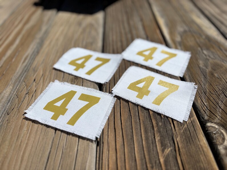May include: Four white fabric patches, each displaying the number 47 in gold. The patches have a frayed edge and are set against a weathered wooden background. The patches are square in shape.