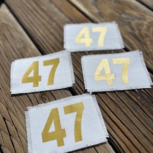May include: Four rectangular white patches, each displaying the number "47" in gold. The patches have frayed edges and are arranged on a wooden surface. The gold numbers are centrally positioned.