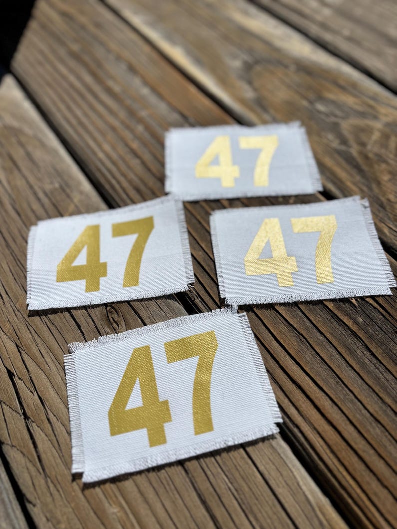 May include: Four rectangular white patches, each displaying the number "47" in gold. The patches have frayed edges and are arranged on a wooden surface. The gold numbers are centrally positioned.