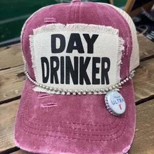 May include: A burgundy and tan trucker hat with the words "DAY DRINKER" in black block letters on a white patch. The hat has a silver beaded trim and a bottle cap attached. The hat is distressed.