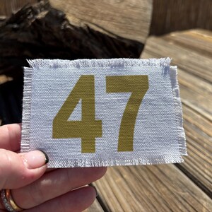 May include: A rectangular, light grey fabric patch with frayed edges. The number "47" is printed in a gold colour. The patch is held up by a hand.