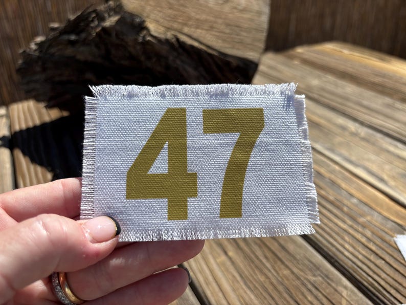May include: A rectangular, light grey fabric patch with frayed edges. The number "47" is printed in a gold colour. The patch is held up by a hand.