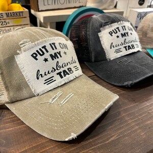 May include: Three distressed baseball caps with a mesh back. The caps are beige, black, and turquoise. Each cap has a white patch with black text that reads "Put it on my husbands tab".