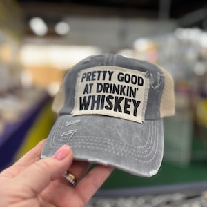 Puede incluir: Una gorra de béisbol gris y beige con un aspecto desgastado. La gorra tiene un parche en la parte delantera que dice "Pretty Good At Drinkin' Whiskey".