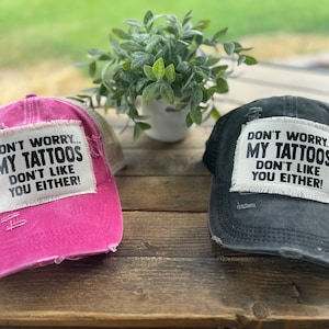 May include: Two distressed trucker hats with a white patch that reads "Don't worry... My tattoos don't like you either!" One hat is pink and the other is black.