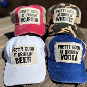 May include: Four distressed trucker hats with the text "PRETTY GOOD AT DRINKIN'" followed by "BOURBON", "WHISKEY", "BEER", and "VODKA". The hats are in red, camouflage, white, and blue, with tan mesh backs.
