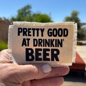 May include: A rectangular, beige fabric patch with frayed edges. Bold black text reads "PRETTY GOOD AT DRINKIN' BEER". The patch is held up against a sunny outdoor background.