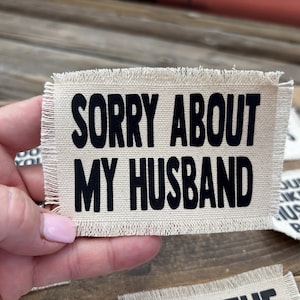 May include: A rectangular patch with frayed edges, made of beige fabric. Bold black text reads "SORRY ABOUT MY HUSBAND". The patch is held up against a wooden surface, with other similar patches visible in the background.