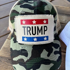 Może przedstawiać: Czapka bejsbolowa w kamuflażu z siatkowym tyłem i postarzaną naszywką. Na naszywce widnieje napis "TRUMP" czarnymi literami, z czerwonymi i niebieskimi paskami oraz białymi gwiazdami.