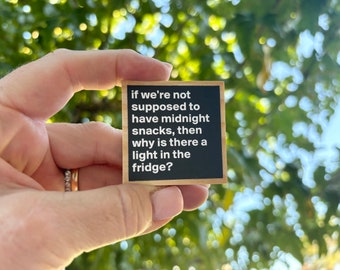 May include: A small, square sign with a black background and a wooden frame. The sign features white text that reads, "if we're not supposed to have midnight snacks, then why is there a light in the fridge?"