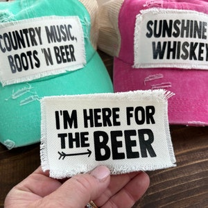 May include: Three trucker hats with different patches. The first hat is turquoise with a white patch that says "COUNTRY MUSIC, BOOTS 'N BEER". The second hat is pink with a white patch that says "SUNSHINE & WHISKEY". The third hat is white with a white patch that says "I'M HERE FOR THE BEER" with an arrow pointing to the right.