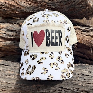 Może przedstawiać: Biało-beżowa czapka typu trucker z wzorem w cętki. Czapka posiada naszywkę z napisem "I ❤️ BEER" czarnymi literami i czerwonym sercem. Ma siatkową tylną część i zakrzywiony daszek.