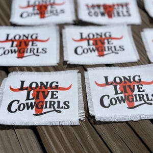 May include: Multiple rectangular white fabric patches with frayed edges. Each patch features the black text "LONG LIVE COWGIRLS" printed over a stylized red graphic of a longhorn steer skull. The design is centered on each individual fabric piece.