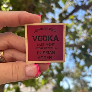 May include: A square wooden sign with a dark red center and light wood border. The text in black reads: I DRANK SO MUCH VODKA LAST NIGHT I WOKE UP WITH A RUSSIAN ACCENT. The sign is held against a blurred outdoor background of green leaves and blue sky.