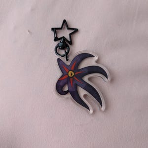 May include: A clear acrylic keyring featuring a stylized, dark purple and black creature with a red and yellow eye. The keyring has a black star-shaped clasp.