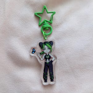 May include: A clear acrylic keychain featuring a cartoon character with green hair and a black and green outfit, holding a blue butterfly. The keychain has a green star-shaped clip.