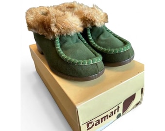 May include: A pair of dark green slippers with tan faux fur trim around the collar and a thick stitched seam along the top. The slippers are resting on a tan cardboard box with the brand name Damart printed in dark red and black text.