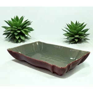 May include: A rectangular ceramic serving dish with a scalloped edge. The interior is a muted green, and the exterior edge is a deep burgundy. Two green succulent plants are in the background.