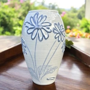 May include: A white ceramic vase with a rounded body and a narrow opening. The exterior features raised, embossed blue floral designs depicting daisies with long stems and leaves, creating a textured surface across the front of the vessel.