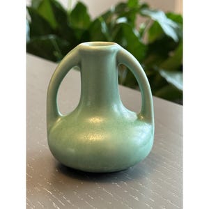 May include: A small, matte, sage green ceramic vase with a rounded base and two symmetrical, arched handles extending from the neck to the shoulder. The surface has a smooth, uniform finish with subtle variations in the light green color tone.