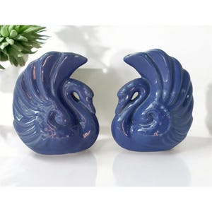 May include: Two matching blue ceramic swan figurines. The swans have detailed wing and feather designs, with curved necks and a glossy finish. The figurines are displayed on a white surface.